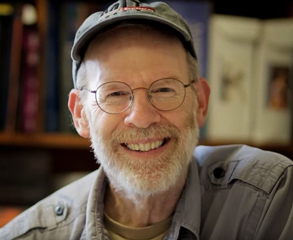 Smiling older man wearing glasses, cap, and khaki shirt in indoor setting