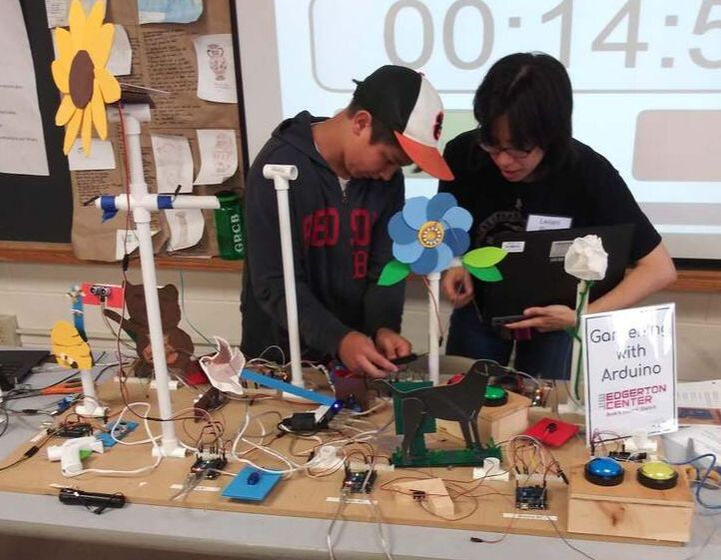 Two students examining Arduino robotics project with flower mechanism on classroom table display.