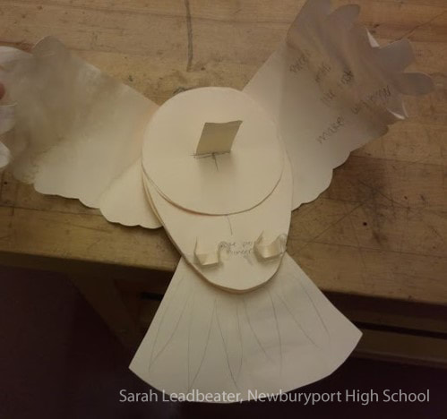 White paper angel craft with smiling face, wings, and pleated dress on wood.