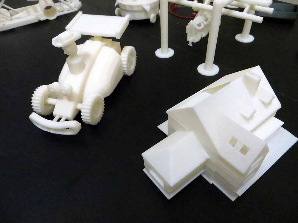 White 3D printed parts including toy car, house model, and mechanical components on dark surface.