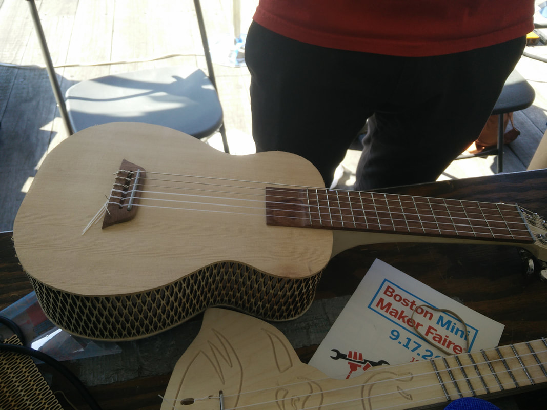 Person holding handmade wooden acoustic guitar at Boston Mint Maker Faire outdoor event