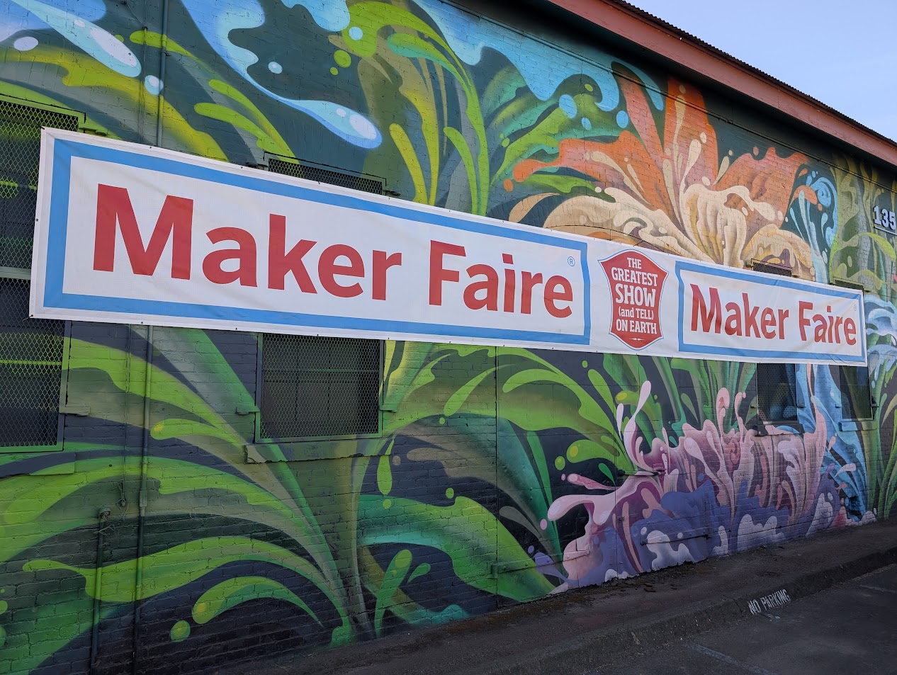 A banner hung on a brightly painted brick wall reads "Maker Faire The Greatest Show (and Tell) on Earth.