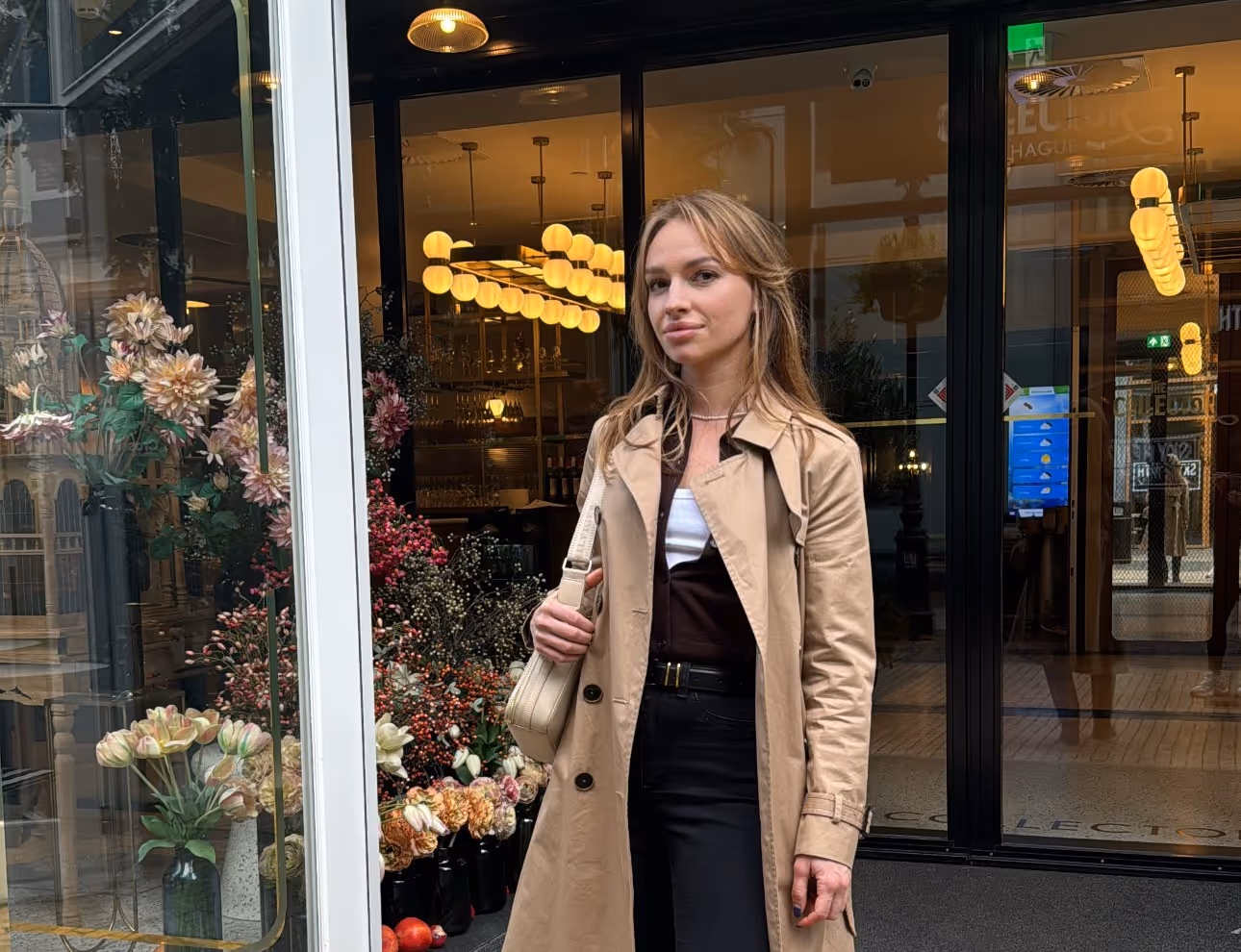 Young woman in beige trench coat carrying a beige bag stands outside a flower shop window display.