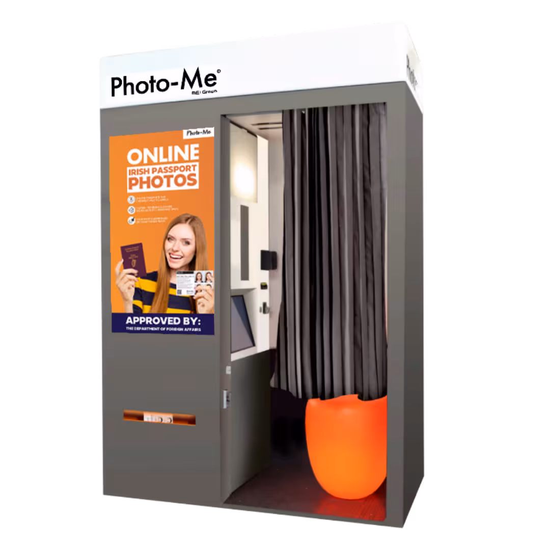 Photo-Me booth for online Irish passport photos featuring a smiling woman holding a passport and photo card, with a black curtain and orange seat inside.