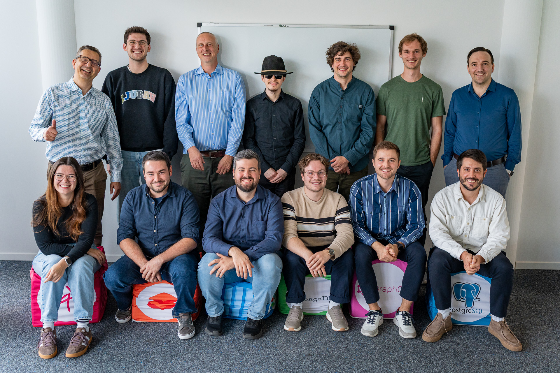 Drei Personen in Schürzen halten gemeinsam einen großen Käselaib in einer traditionellen Käserei. Im Hintergrund sind rustikale Werkzeuge und eine dunkle Wand zu sehen.