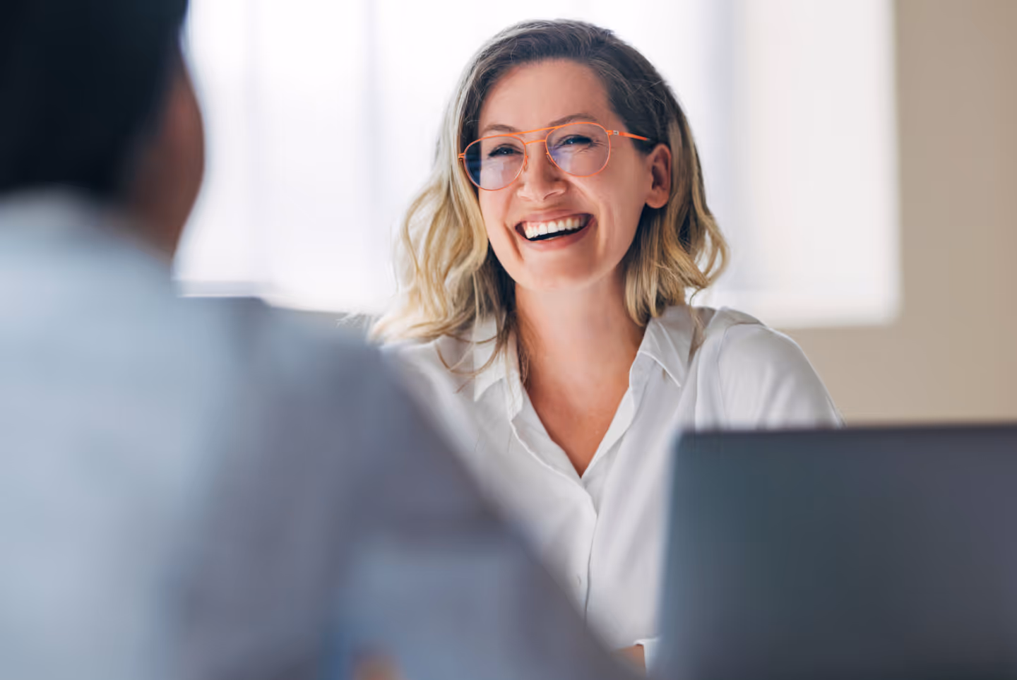 A woman with glasses chatting to someone in a meeting, smiling.