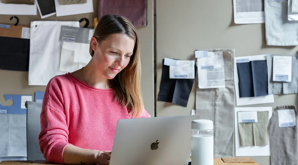 Woman in a pink sweater working on a silver MacBook laptop at a desk with fabric samples on the wall behind her.