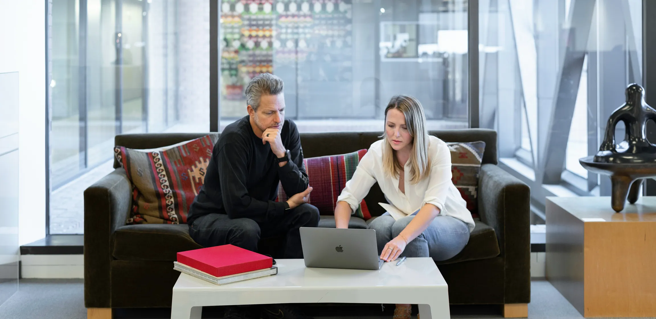 Man and woman sitting on a couch looking at a laptop on a white coffee table in a modern office setting.