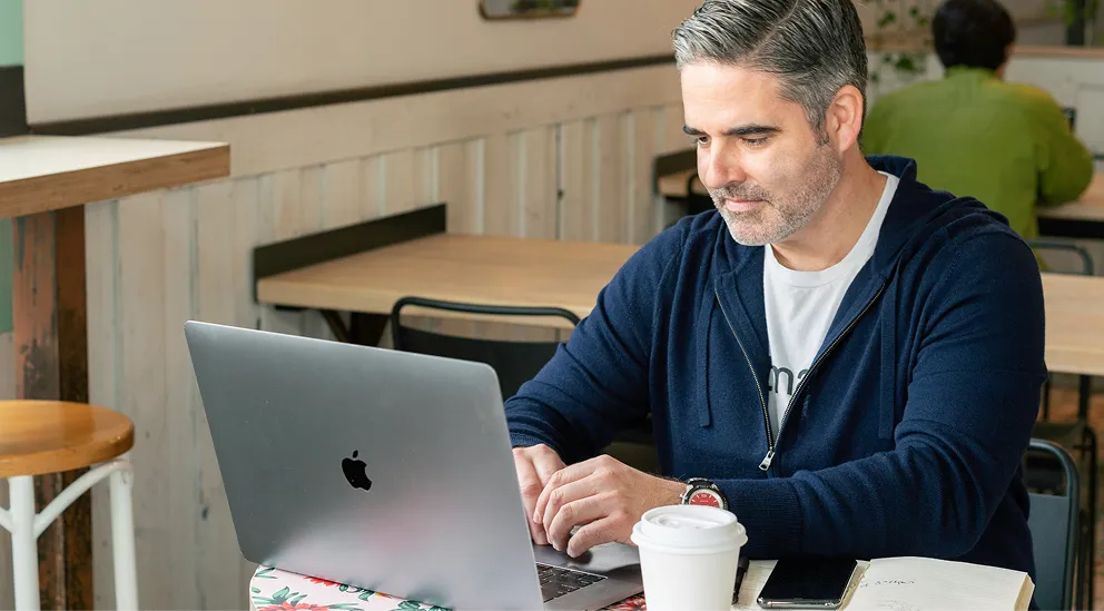 Man with gray hair and beard working on a laptop at a café table with a coffee cup, notebook, and smartphone.