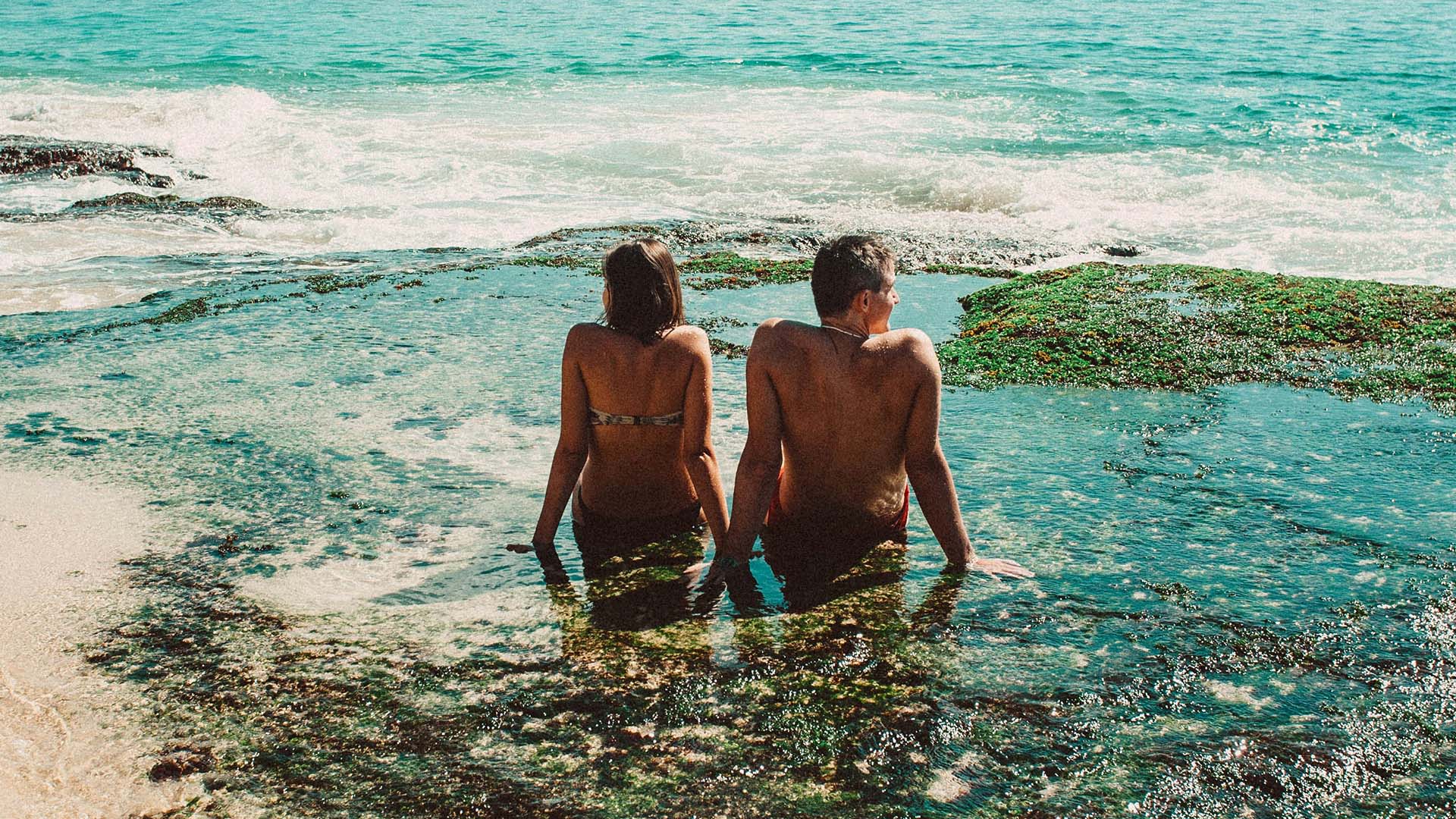 Two people sitting together in shallow sea water on a sunny coastline. Image via Unsplash