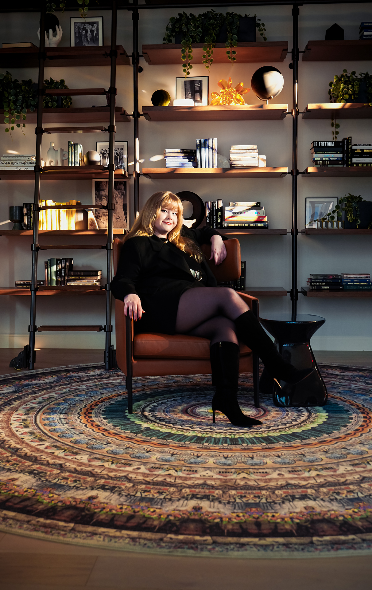 Woman with blonde hair wearing black dress and boots sitting on a brown chair in front of a bookshelf with plants and books.