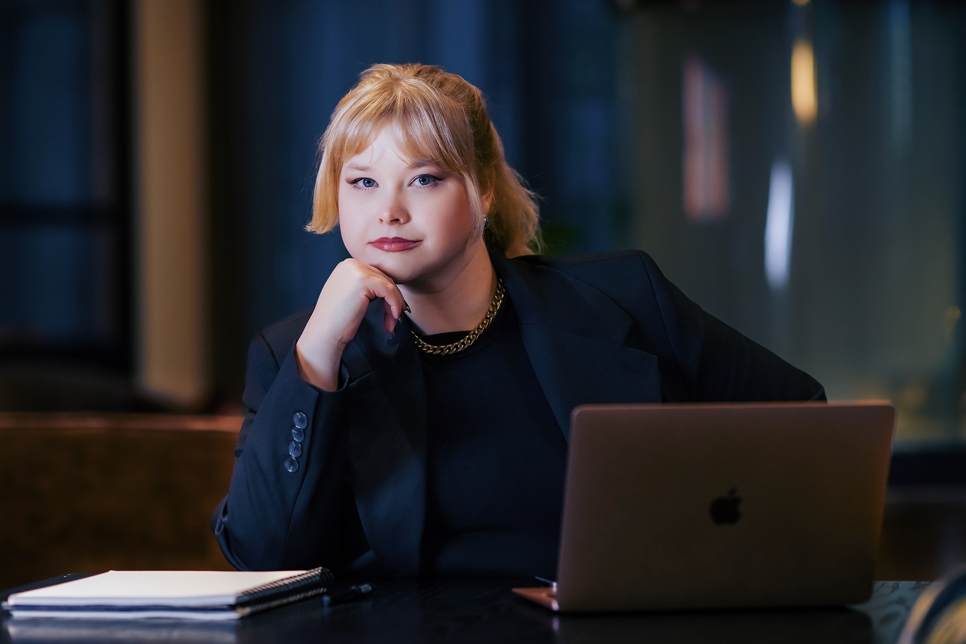 Young woman with blonde hair sitting at a desk with a laptop and notebook, resting her chin on her hand and looking at the camera.