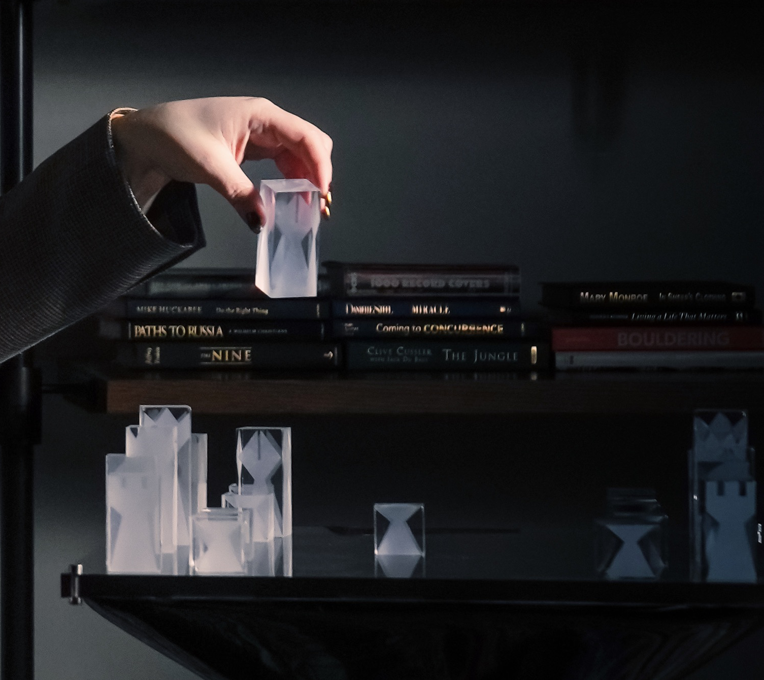 Hand holding a transparent hourglass-shaped glass piece above a black shelf with multiple similar glass pieces and a row of books in the background.