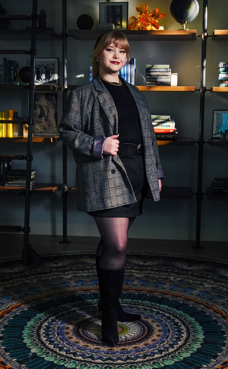 Young woman wearing a gray plaid blazer, black dress, and knee-high boots standing on a colorful patterned rug in front of a bookshelf.