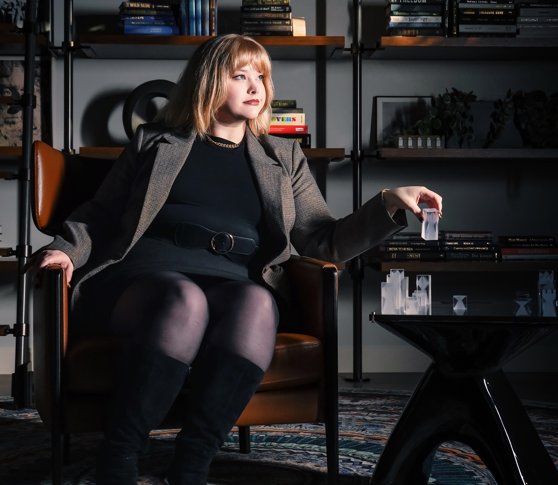 Woman in a black dress and blazer sitting in a leather chair, holding a frosted glass chess piece above a glass chessboard.