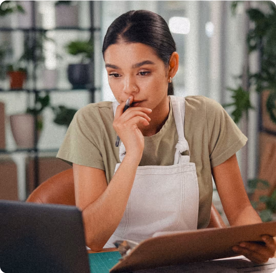 Thoughtful woman in a white apron holding a clipboard and pen, looking at a laptop screen in a workspace with plants in the background.