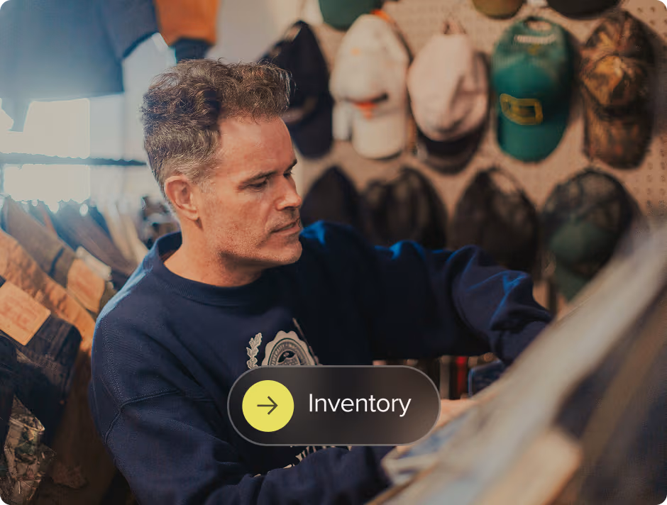 Man wearing a navy sweater browsing clothing items in a store with hats displayed on the wall behind him.