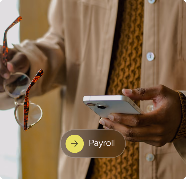 Person holding a smartphone in one hand and eyeglasses in the other, with payroll text overlay.