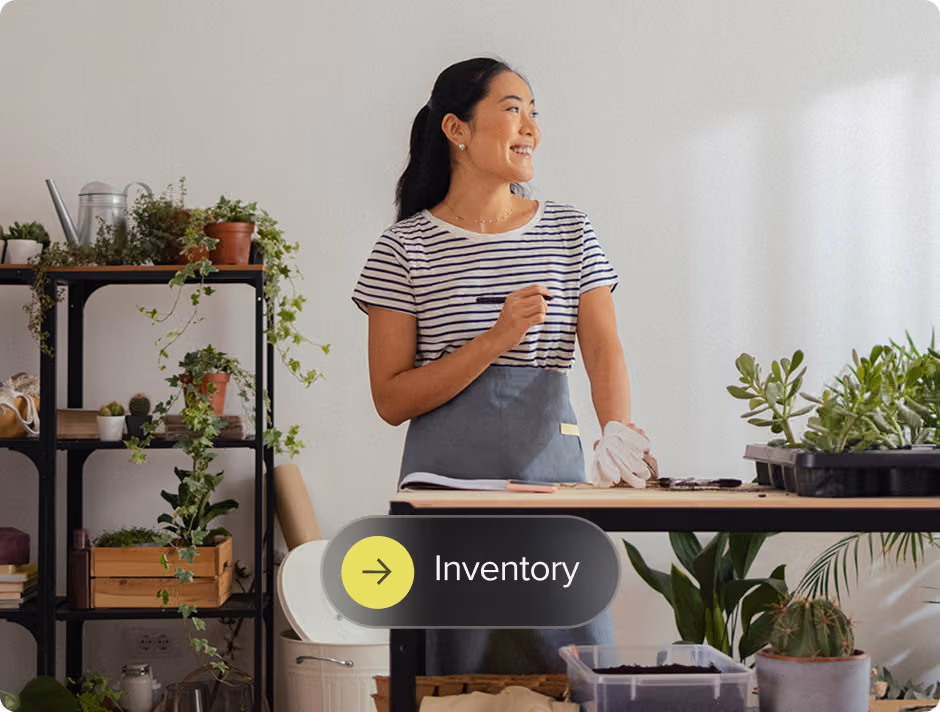 Woman in apron standing at a table with plants and gardening supplies, smiling and looking to the side.