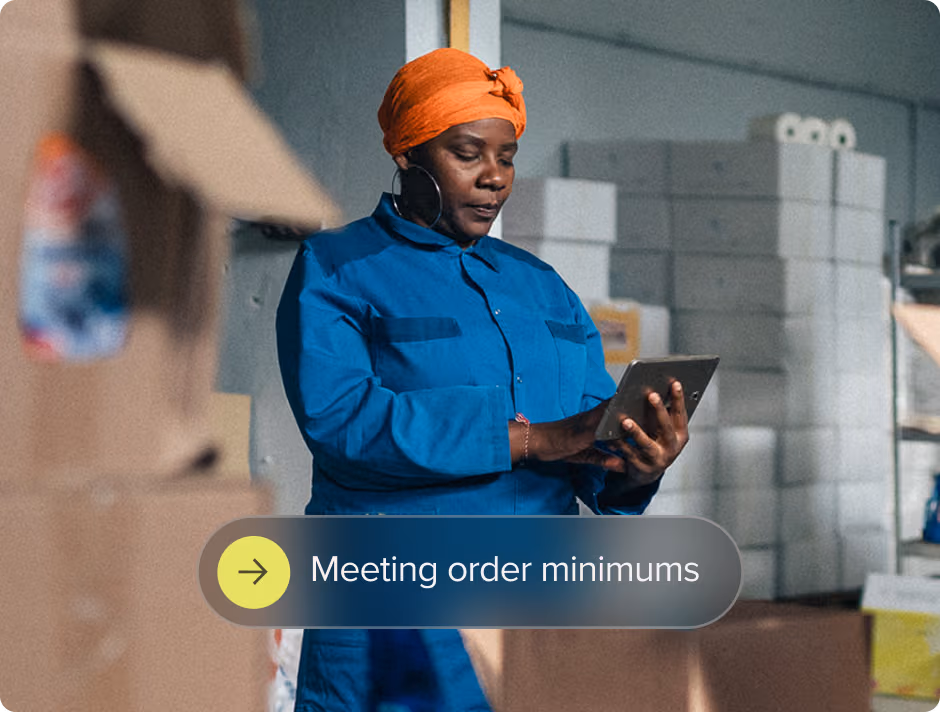 Woman in blue workwear and orange headscarf using a tablet in a warehouse with stacked boxes.