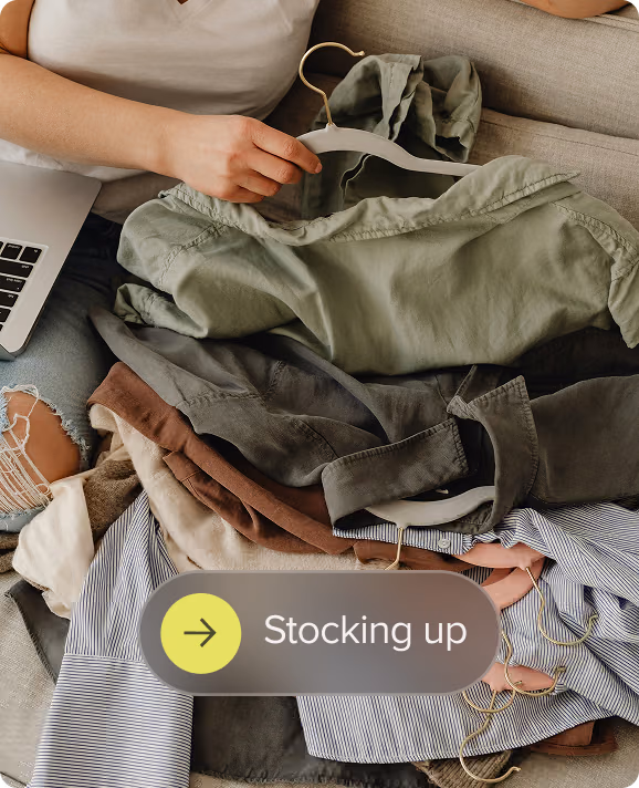 Person holding a green shirt on a hanger over a pile of folded clothes and a laptop beside them.