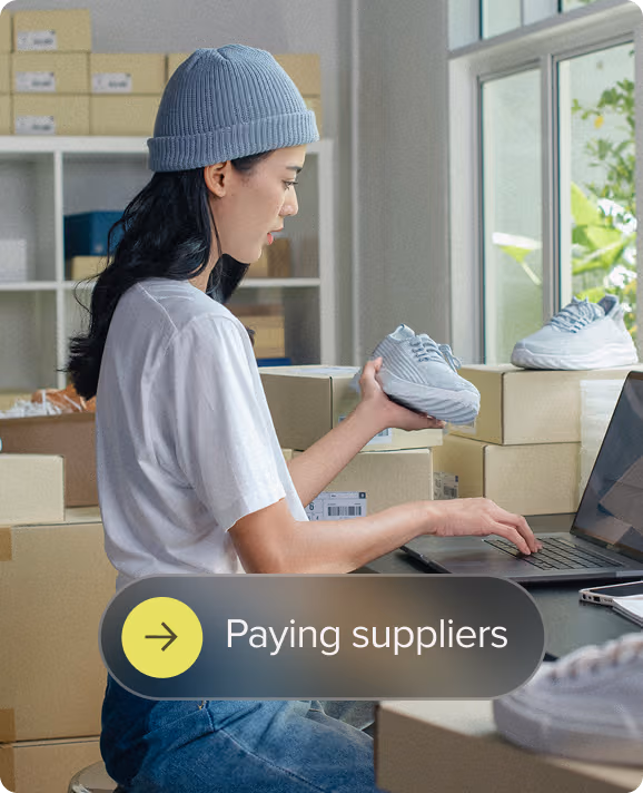 Woman in a beanie holding a sneaker and using a laptop among shipping boxes in a workspace.