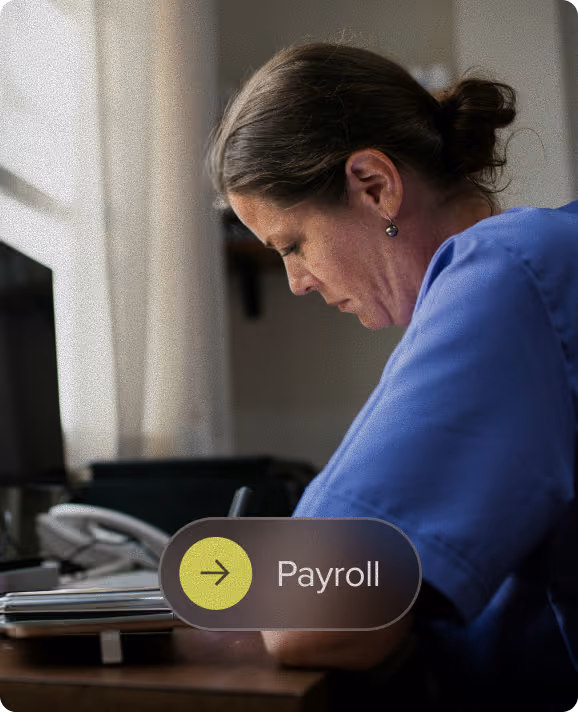 Woman in a blue shirt sitting at a desk, focused on writing or reading, with light coming through a window behind her and a payroll button graphic overlayed at the bottom.