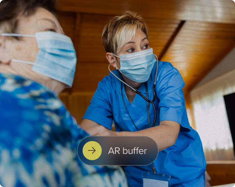 A healthcare worker wearing a blue scrub suit and surgical mask uses a stethoscope to check an elderly patient who is also wearing a mask in a room with wooden walls and ceiling.