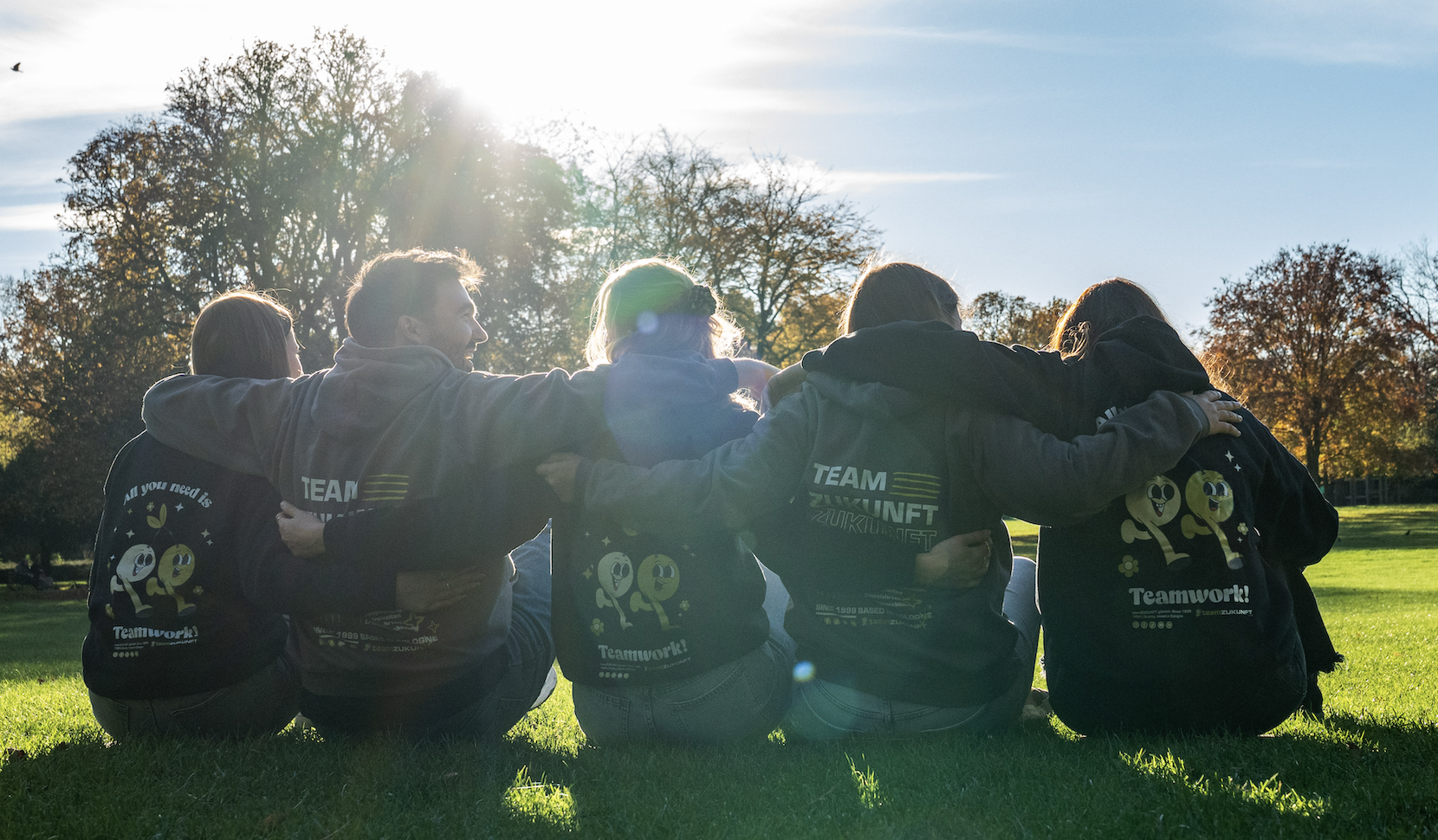 Fünf Personen sitzen im Freien im Gras, die Arme umeinander gelegt, mit Kapuzenpullovern, die Motiv für Teamarbeit zeigen.