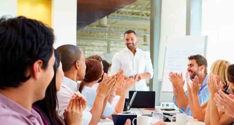 Gruppe von Personen in einem Meetingraum applaudiert einem Mann, der vor einer Flipchart steht.