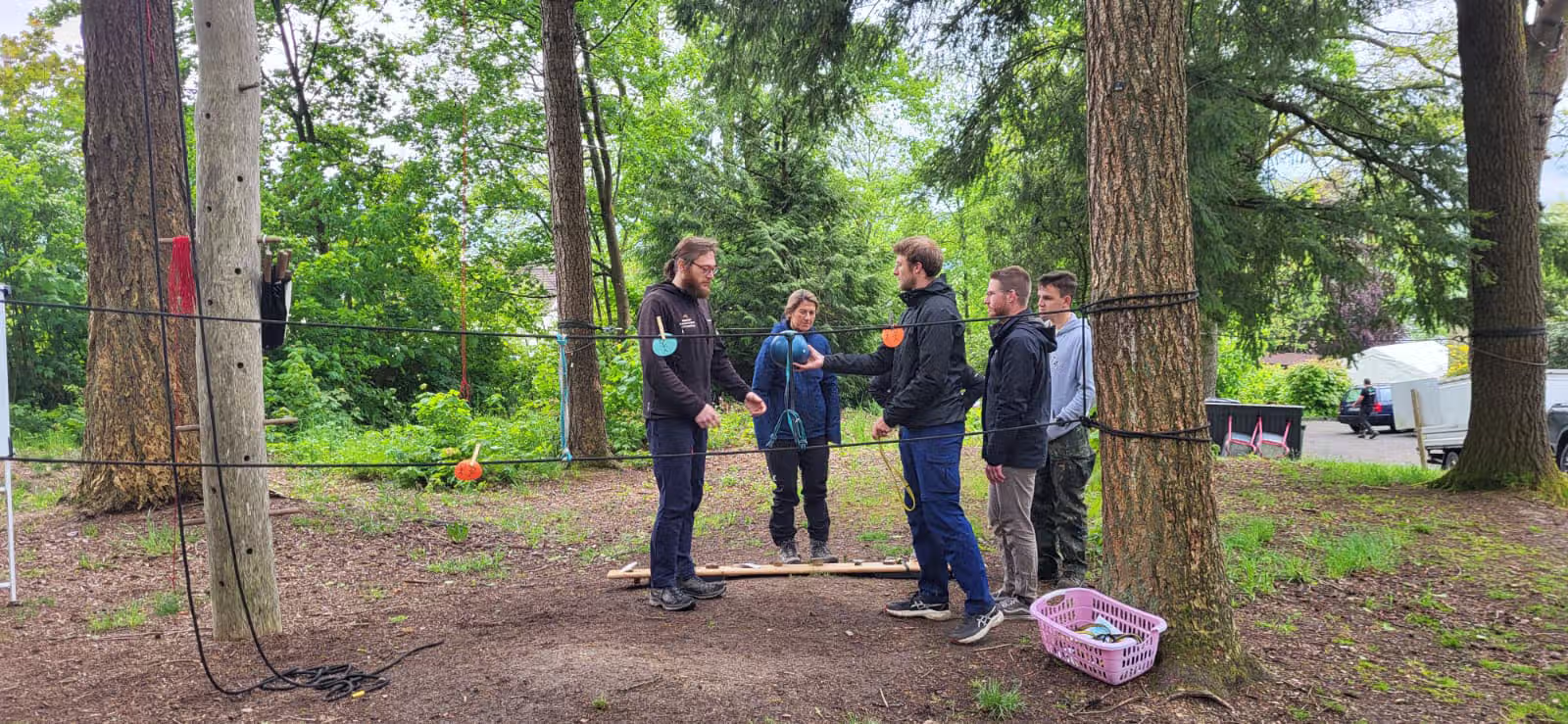 Fünf Personen stehen in einem Waldgebiet auf einem Holzbrett, umgeben von Seilen für Teamtraining.
