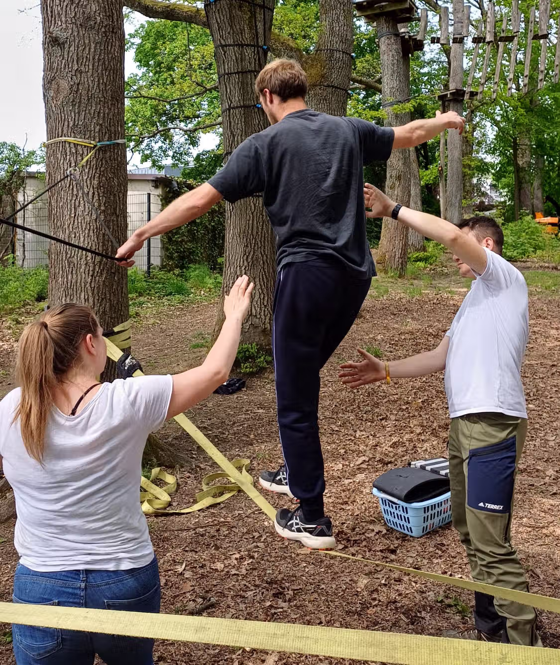 Person balanciert auf Slackline, zwei Menschen helfen beim Stabilisieren im Wald.