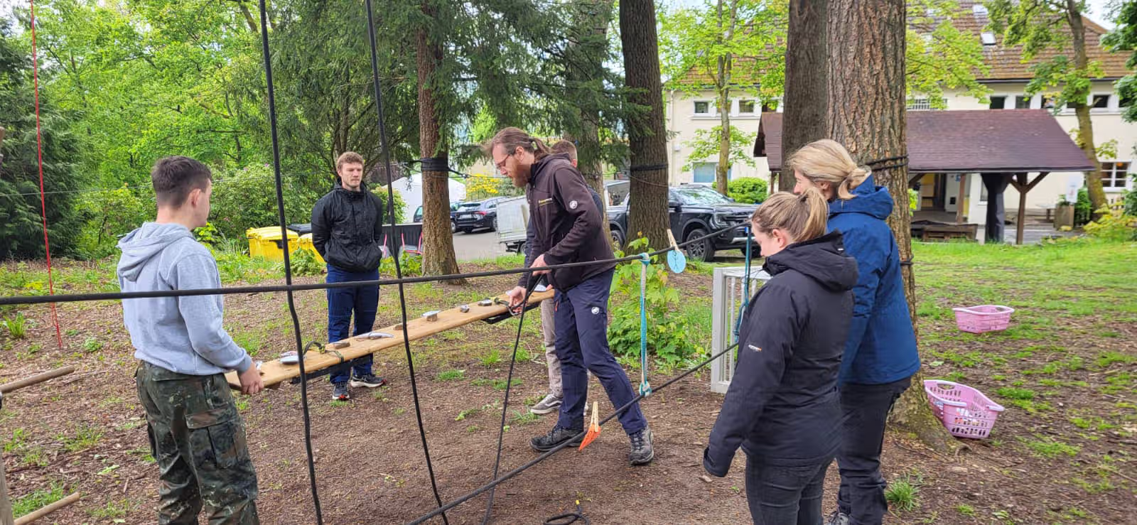 Gruppe von sechs Personen im Wald hält ein Holzbrett mit Steinen, um ein Seilnetz zu überqueren.
