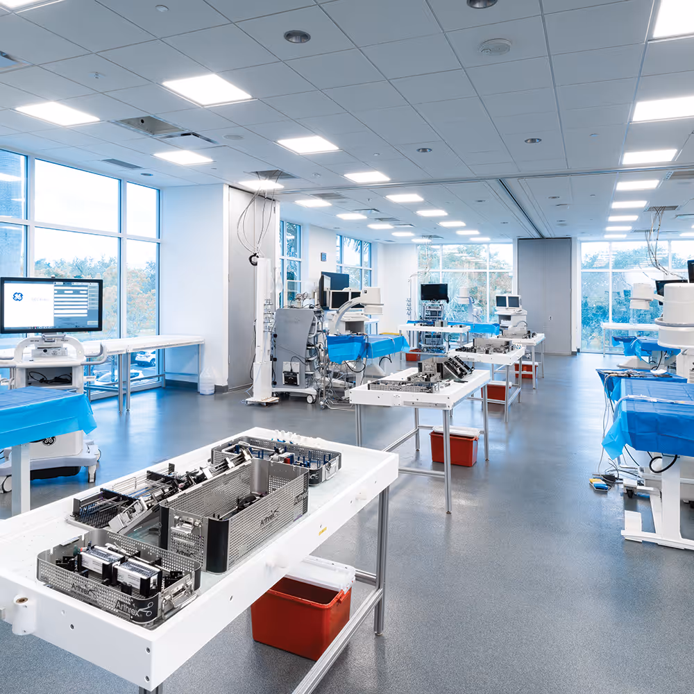 A bright, spacious medical training lab with multiple workstations arranged in rows, each set up with surgical instrument trays and blue drapes. Large windows line the left side of the room, and medical equipment and monitors are positioned throughout the space.
