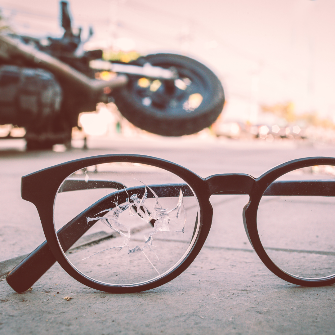 A pair of broken eyeglasses on the road with a crashed motorcycle in the background