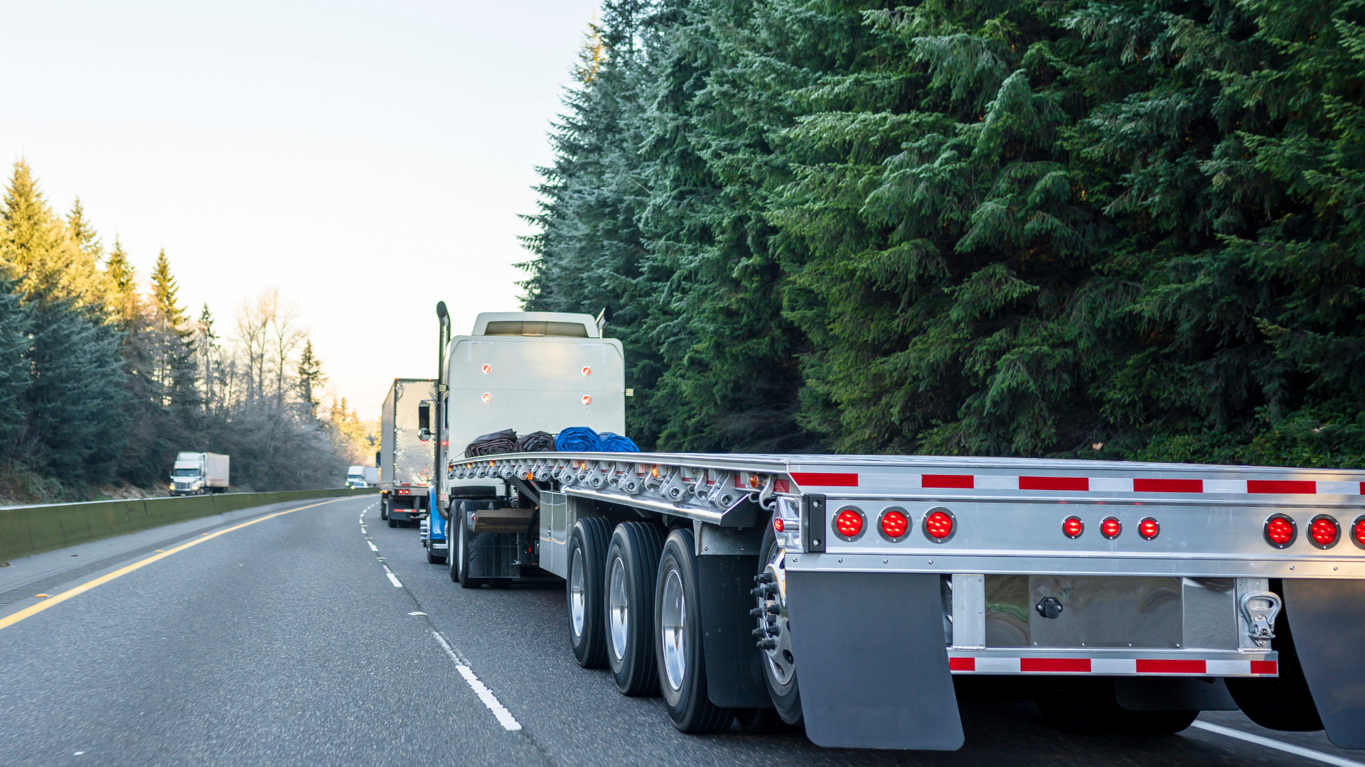 A white semi truck hauling an empty trailer on a freeway surrounded by trees