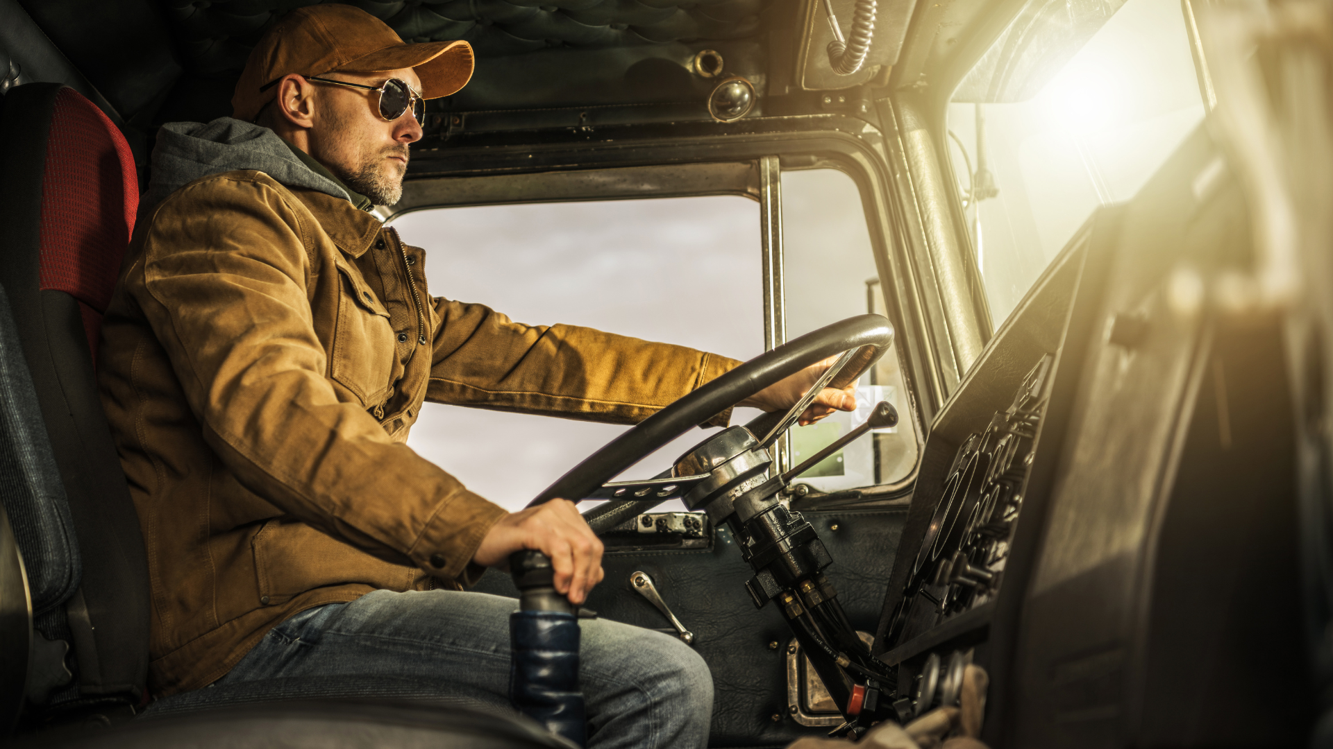 A truck driver wearing a hat, sunglasses and a jacket driving a semi truck