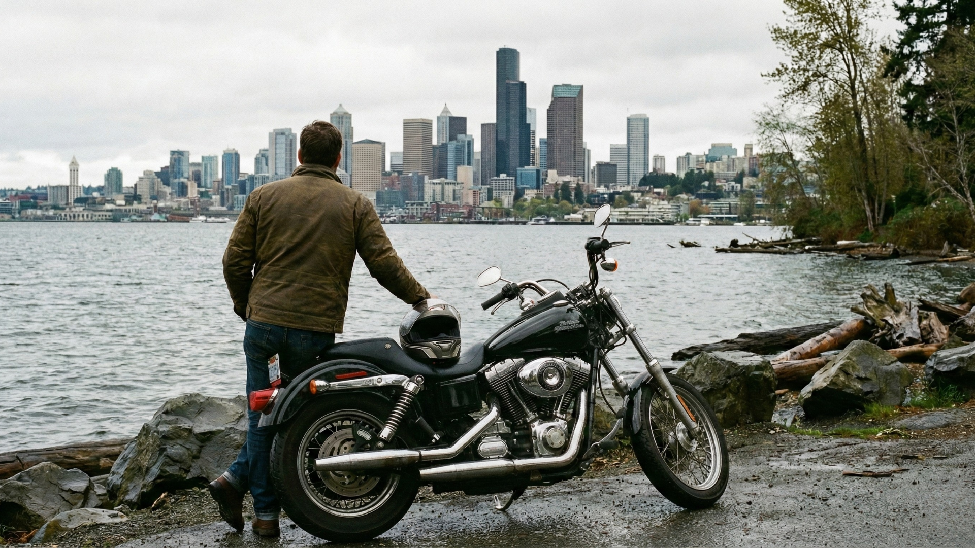 A motorcycle rider standing next to their motorcycle overlooking a lake with a cityscape in the background.
