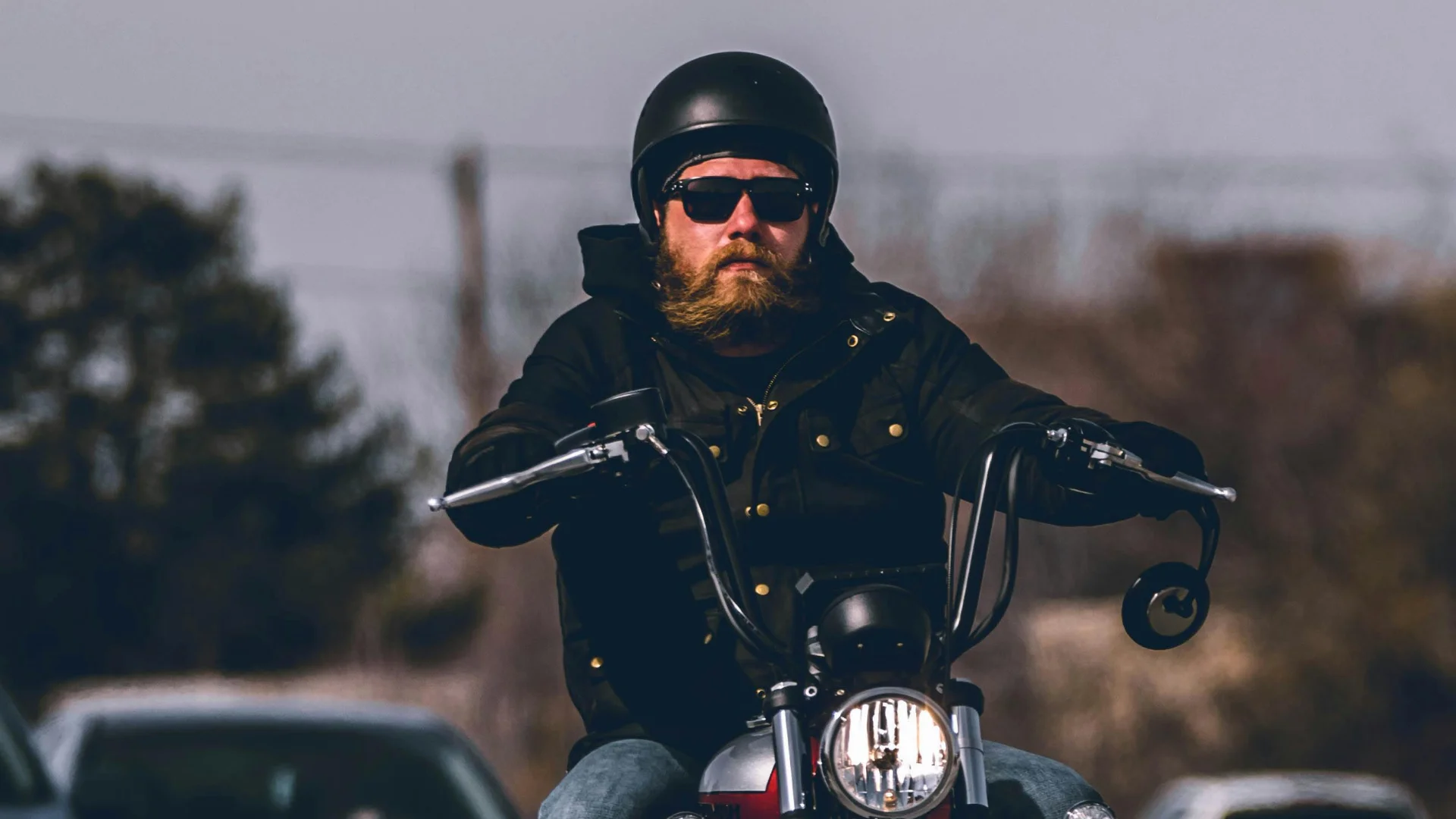 A bearded man with sunglasses and an open faced helmet riding a motorcycel