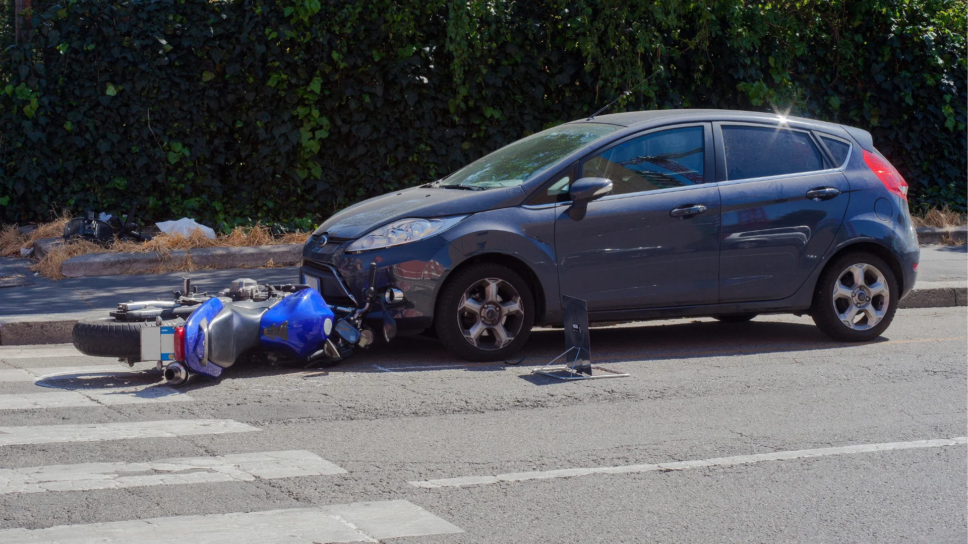 a blue motorcycle on its side on the road that's been hit by a dark blue compact car at an intersection