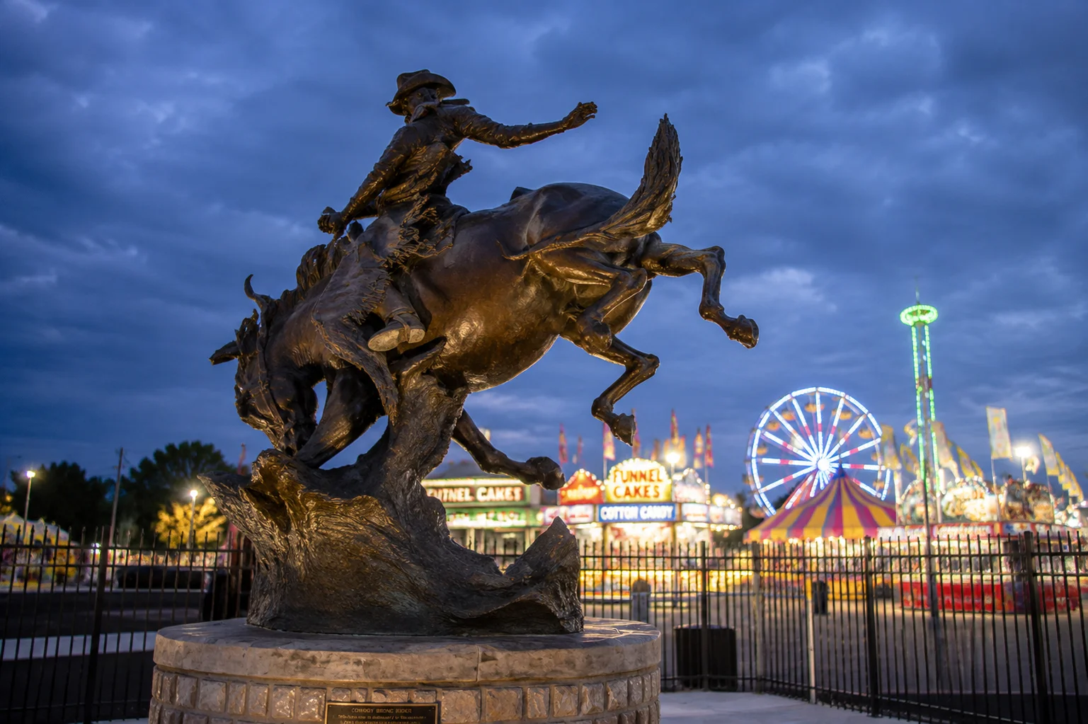 A statue of a cowboy riding a bucking horse with a carnival in the background.