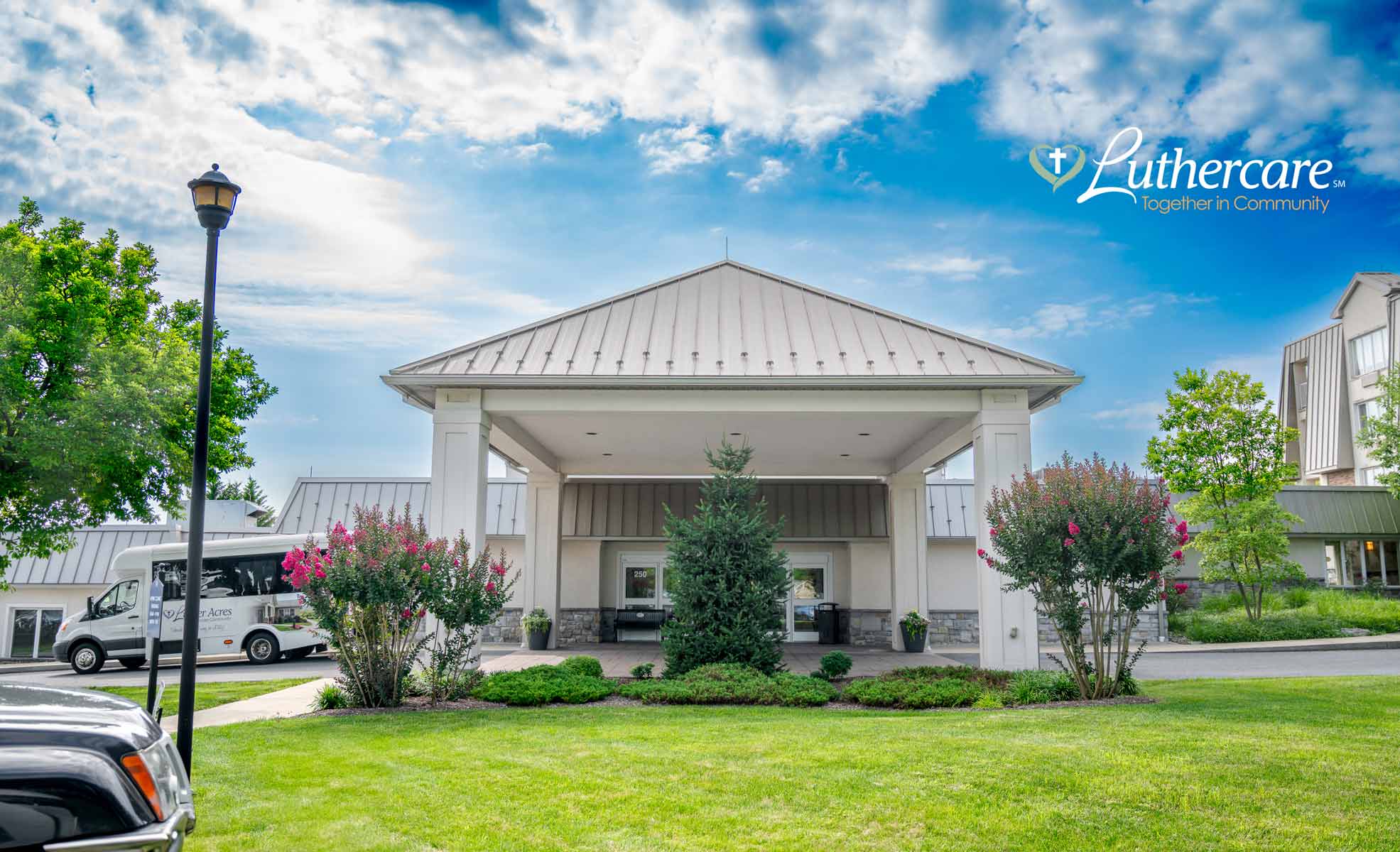 Entrance of a building with a covered portico, surrounded by green lawn, flowering bushes, and trees under a partly cloudy sky.
