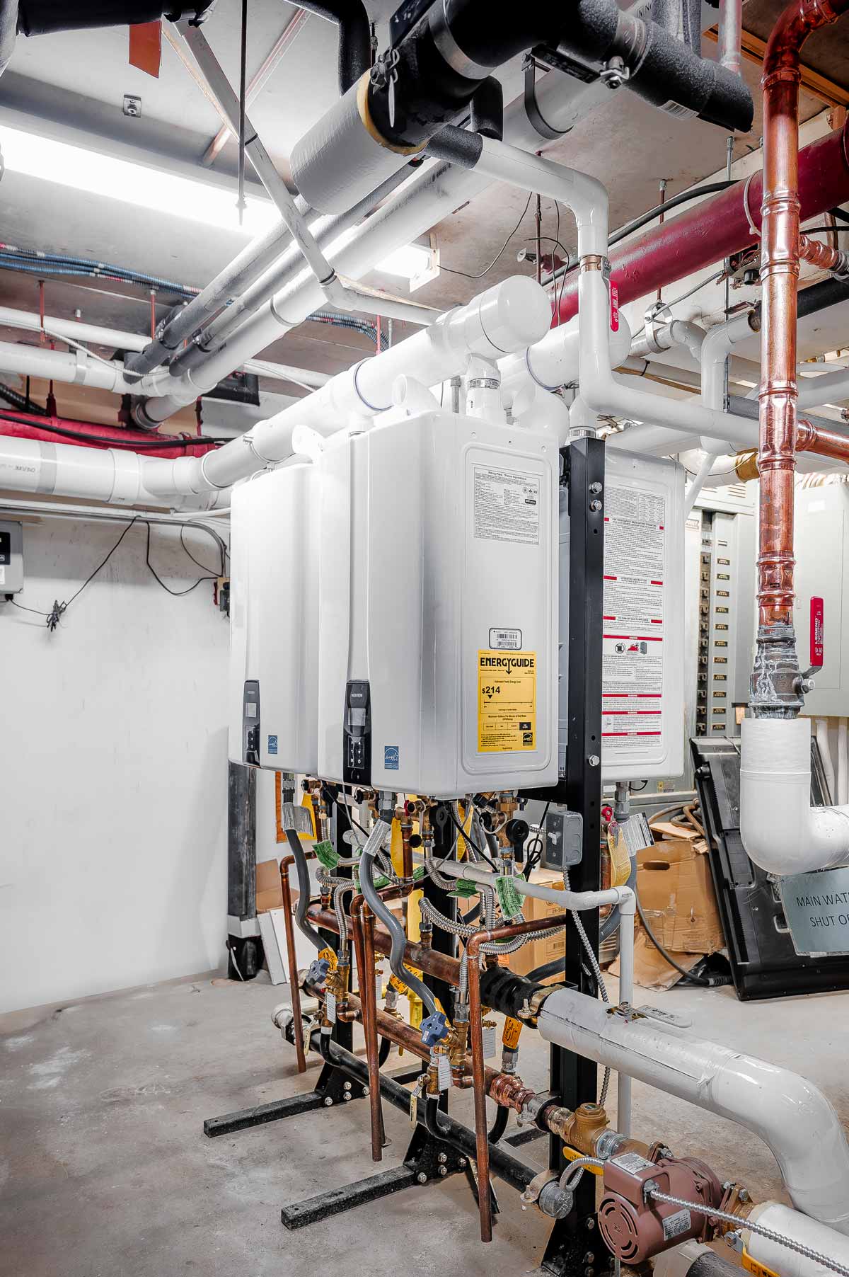 Cluster of modern white tankless water heaters with connected piping and valves in a mechanical room.