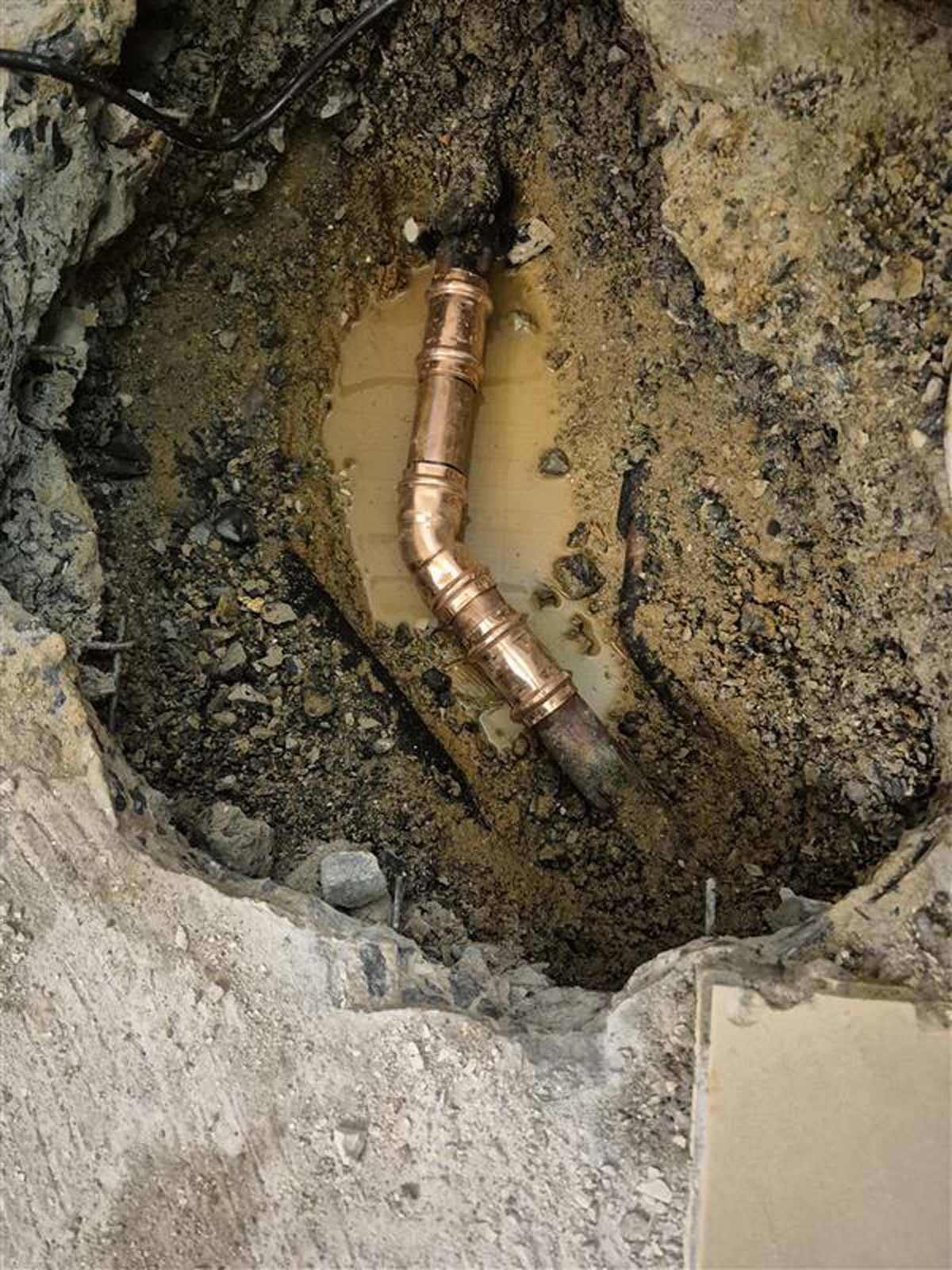Exposed copper plumbing pipe in a muddy excavation surrounded by dirt and concrete edges.