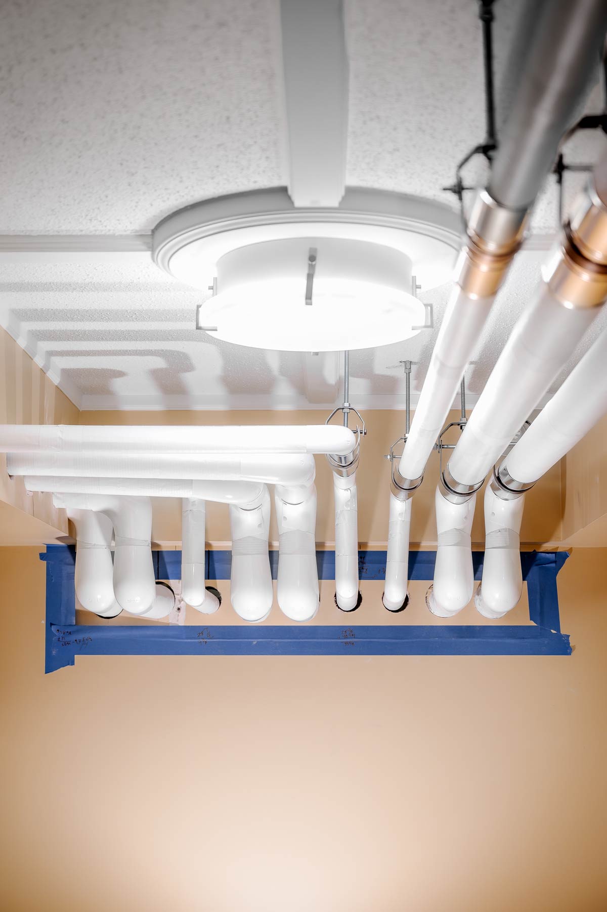 Several white insulated pipes suspended from a ceiling near a round ceiling light, with blue tape framing a rectangular area on the wall.