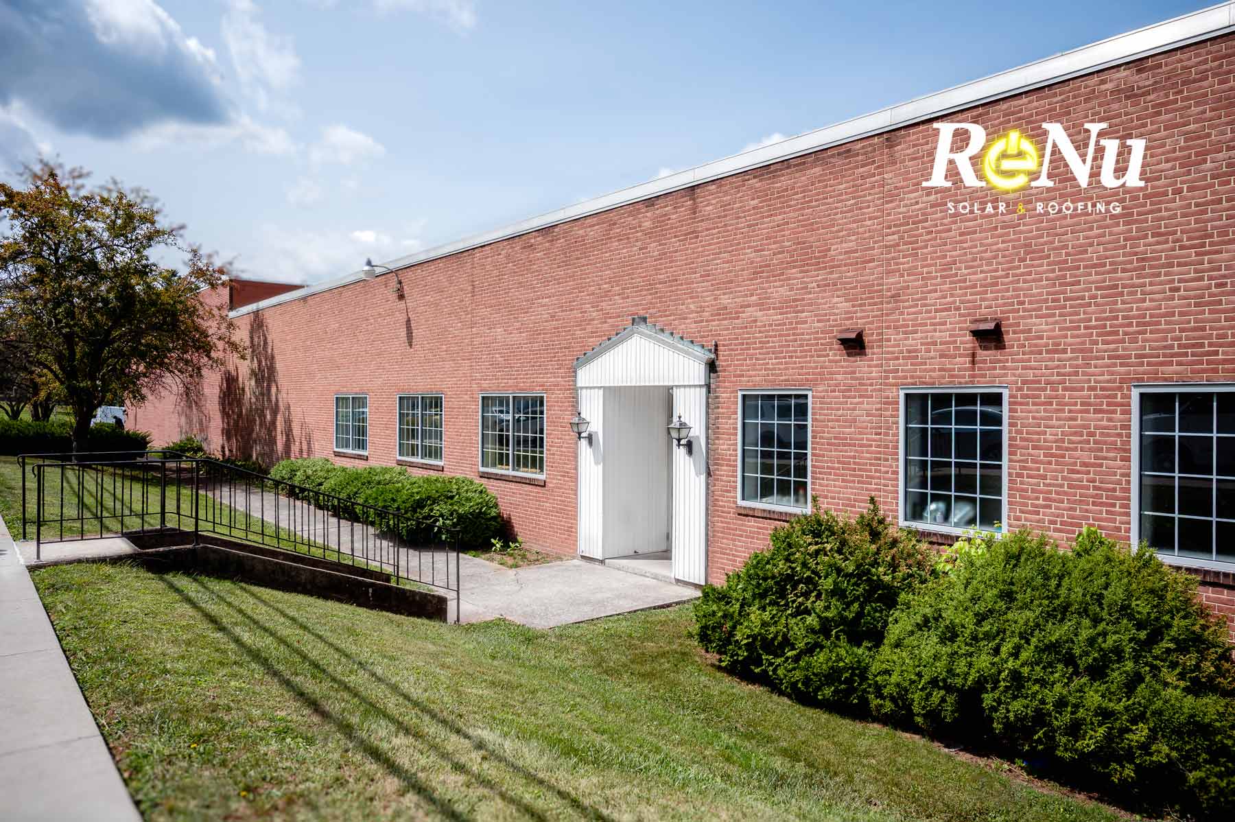 Brick building with white entrance door, several windows, and a manicured lawn with bushes and a tree.