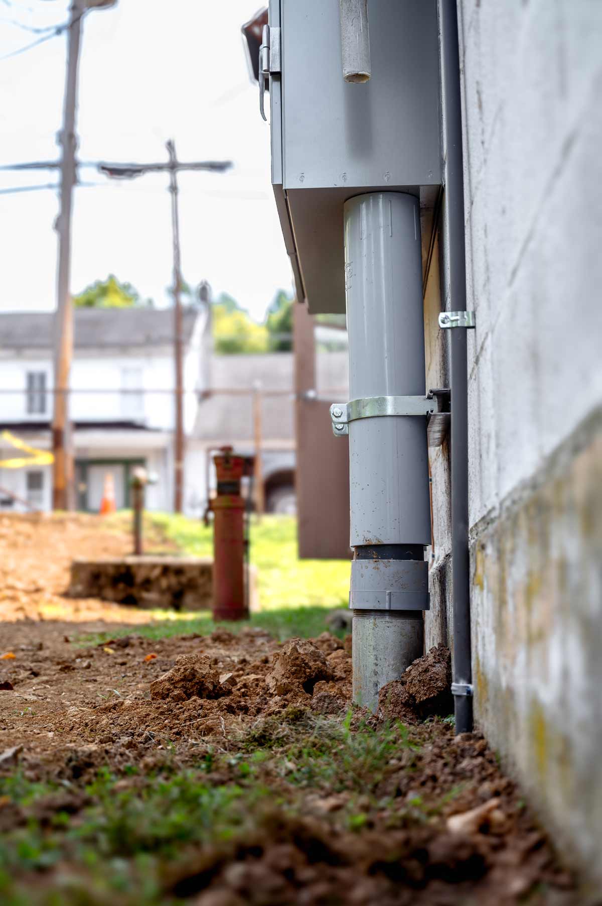 Close-up of a gray electrical conduit attached to the exterior wall of a building with soil and grass in the foreground.