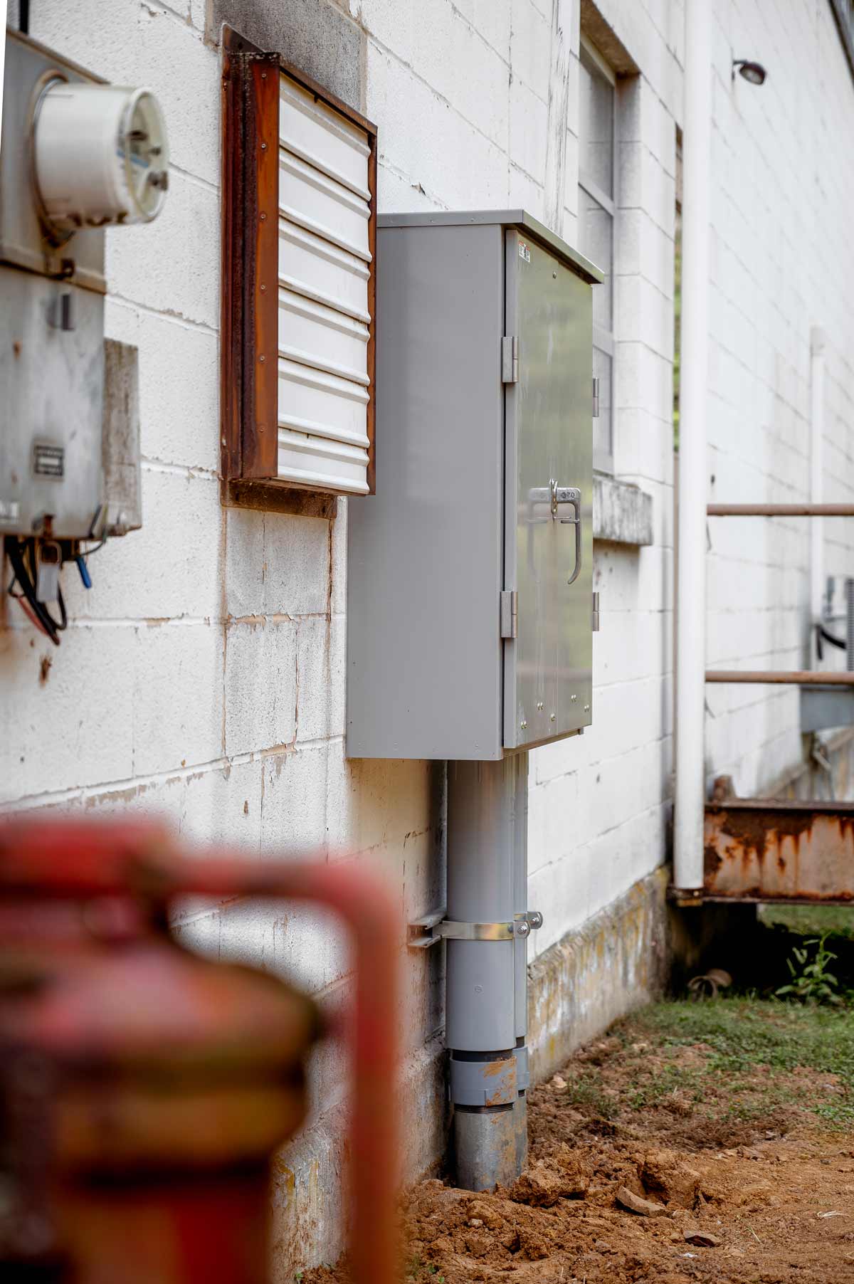 Gray electrical utility box mounted on a white brick wall next to a vent and a rusty pipe, with dirt and grass below.