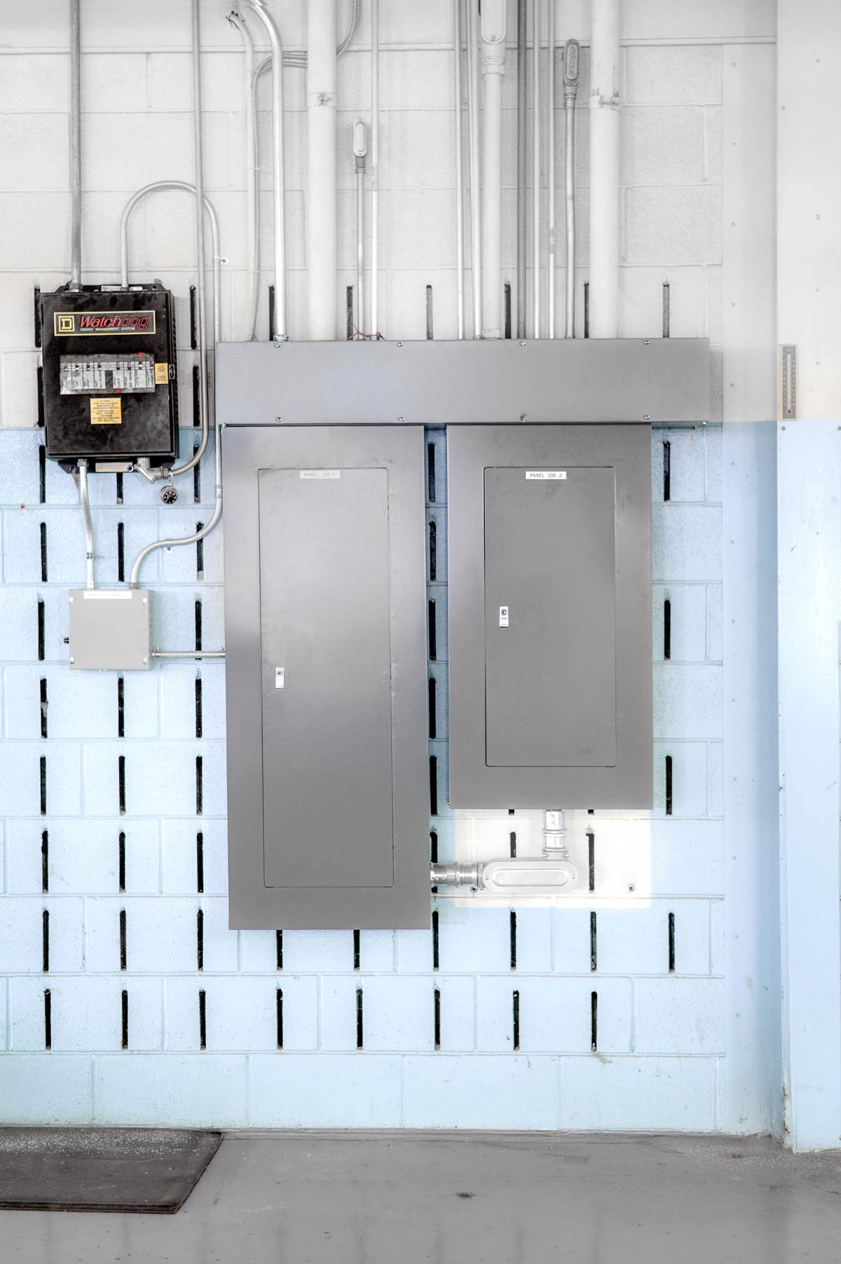 Two gray electrical panels mounted on a light blue and white cement block wall with conduits running above them.