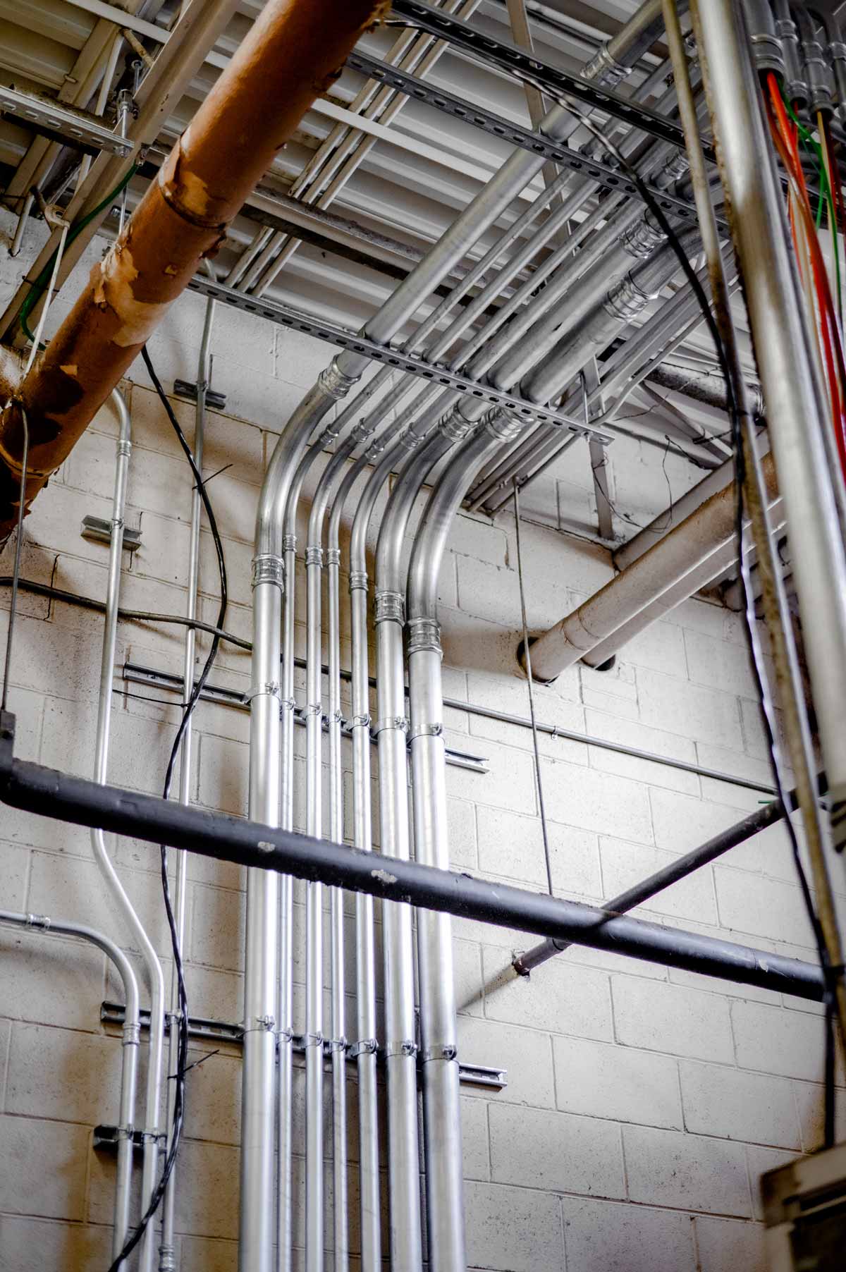 Several silver metal electrical conduit pipes running vertically and bending at the ceiling in an industrial building with white brick walls and overhead pipes.